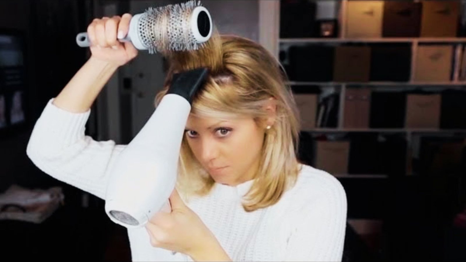 A woman blow-dries her hair, demonstrating a technique for achieving volume while styling Bardot bangs at home.
