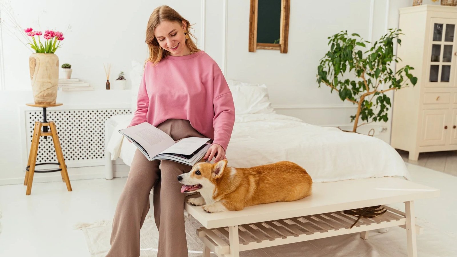 Woman in a cozy pink sweatshirt and beige lounge pants reading a book at home. A corgi dog lies beside her on a bench in a bright, calm bedroom setting.