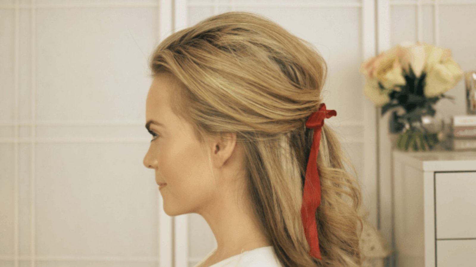 A woman with long blonde hair styled in a Bardot-influenced half-updo, adorned with a red bow.