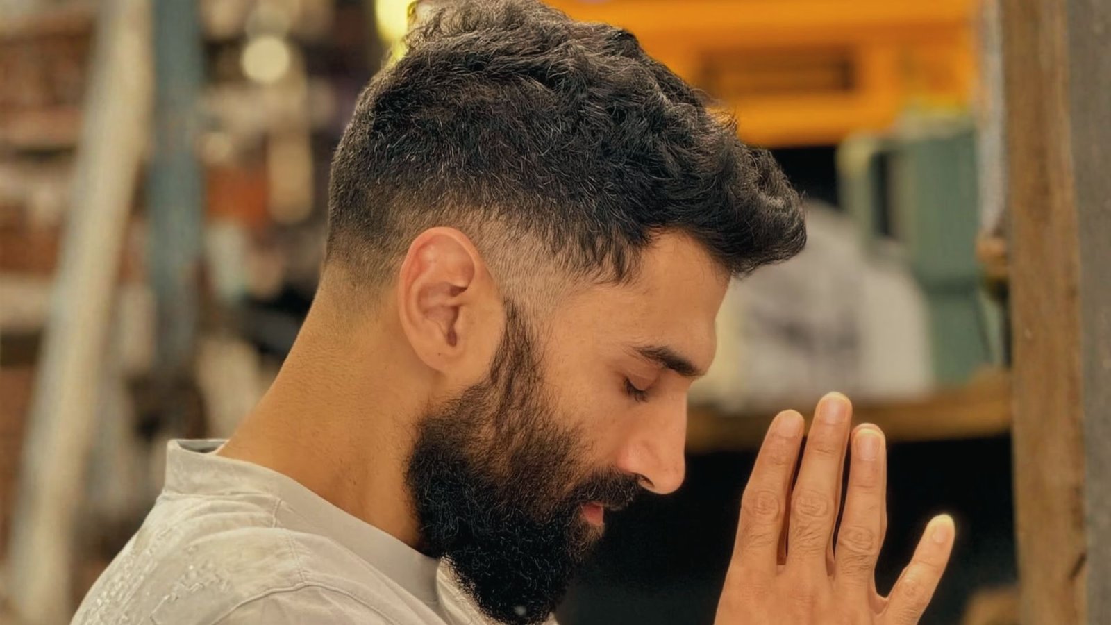 A man with a beard and a quiff disconnected undercut fade is praying with his hands clasped together.