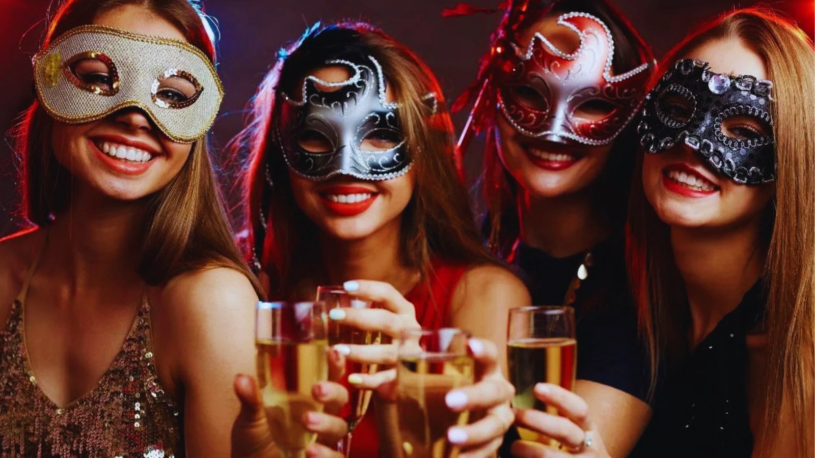 Four women in festive masks smile and raise champagne glasses, dressed in elegant attire at a party, conveying a joyful and celebratory atmosphere.