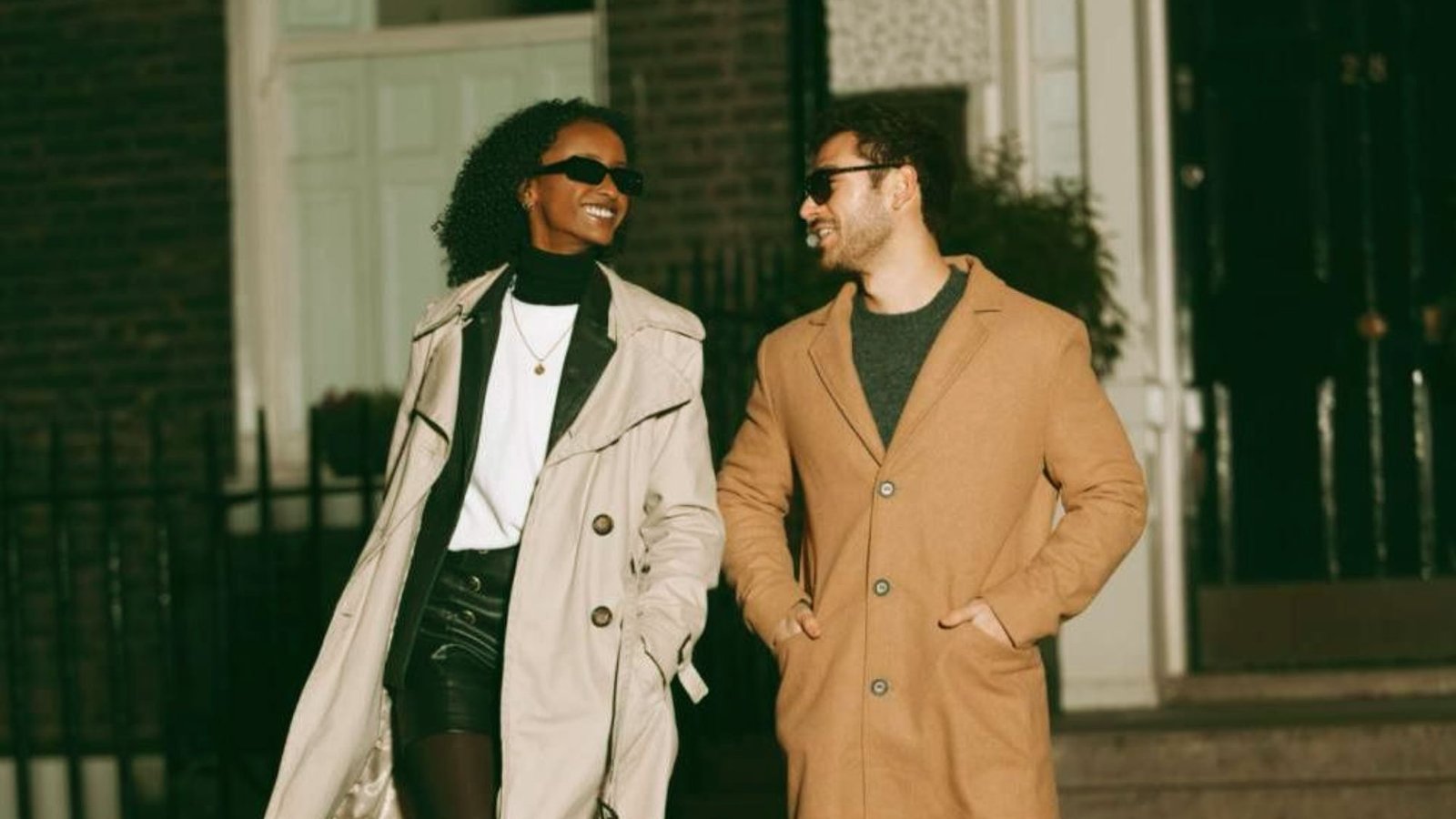 A woman and man walk side by side, both smiling. She's in a beige trench coat, he's in a tan coat. Both wear sunglasses, exuding a cheerful vibe.
