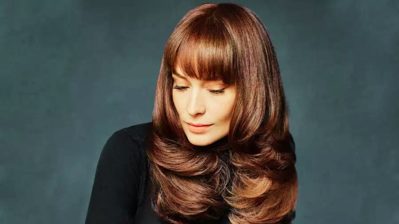 A woman with long brown hair and long diagonal bangs, wearing a black top, poses for the camera.
