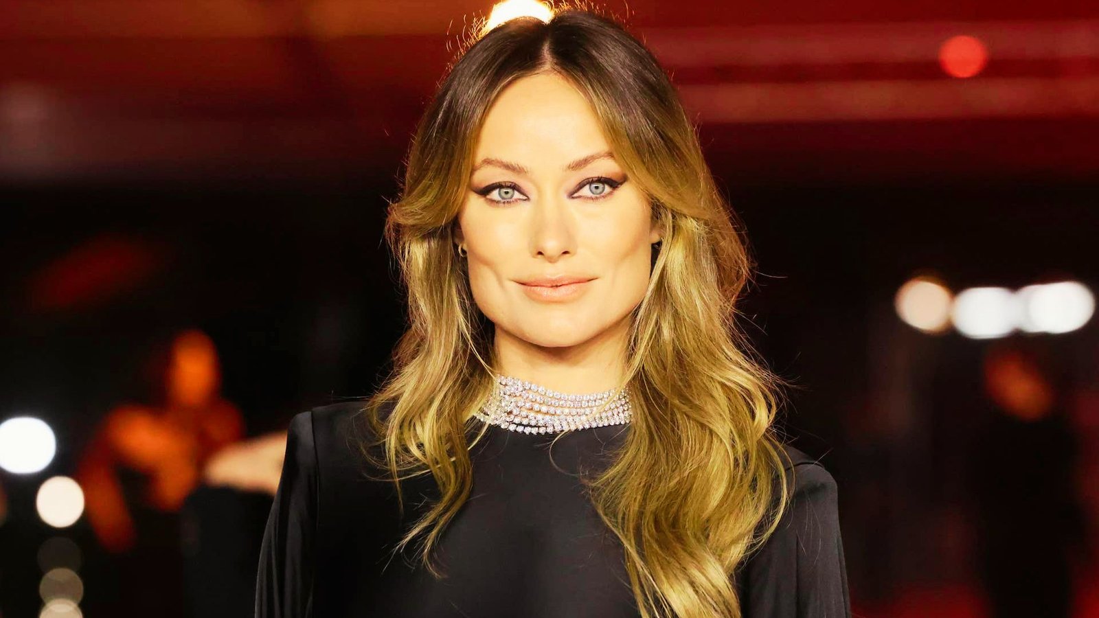 A woman in a black dress stands on a red carpet, showcasing her glass hair finish with long layers and curtain bangs.
