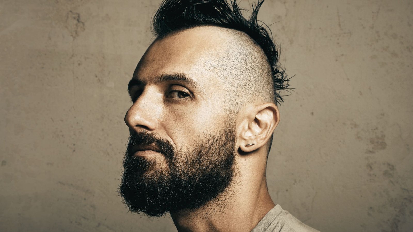 A man with a beard and a mohawk disconnected undercut fade gazes thoughtfully to the side.