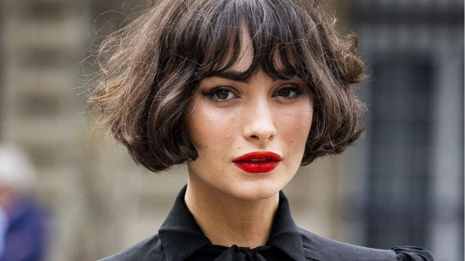 Close-up of a woman with a wavy brunette French bob featuring wispy bangs and red lipstick.