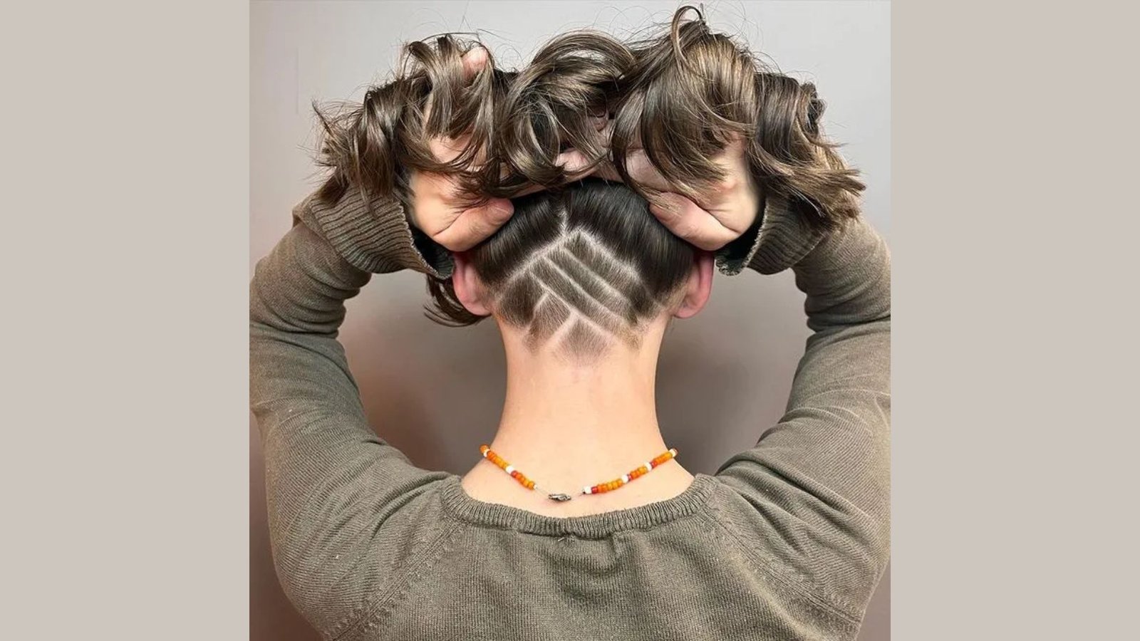 Back view of a person lifting wavy brown hair to reveal a geometric diamond-pattern design shaved into the undercut at the nape.