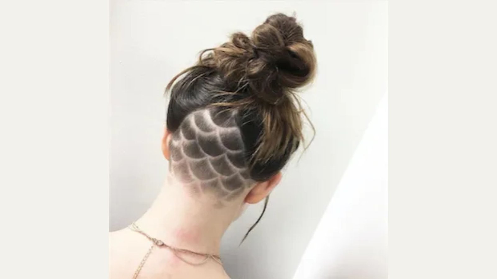 Back view of a woman with a messy topknot revealing a detailed fish-scale pattern shaved into the nape undercut.