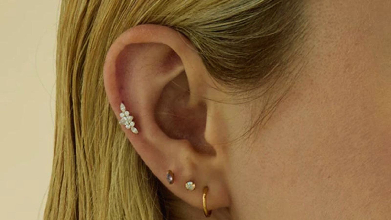 A close-up of a person's ear with multiple piercings. The ear features a curved cluster of small, sparkling gems and three earrings: a small stud, a gem, and a thin gold hoop, set against blonde hair.