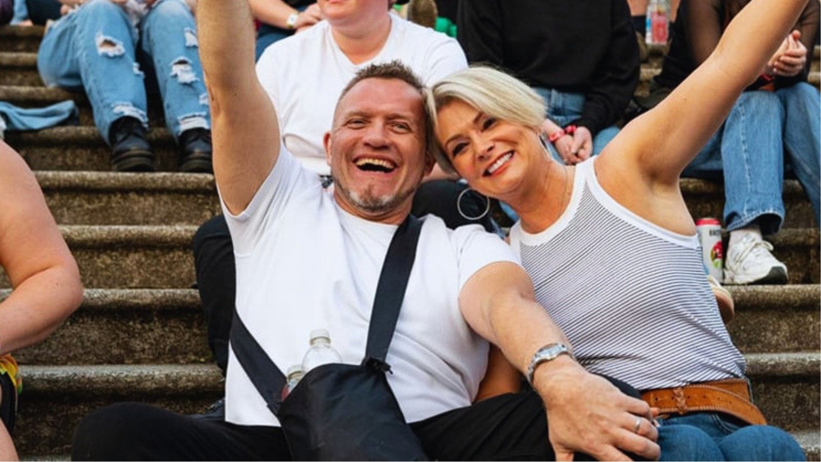 A smiling man and woman sit on stone steps surrounded by people. Both have an arm raised in celebration, conveying a joyful and relaxed atmosphere.