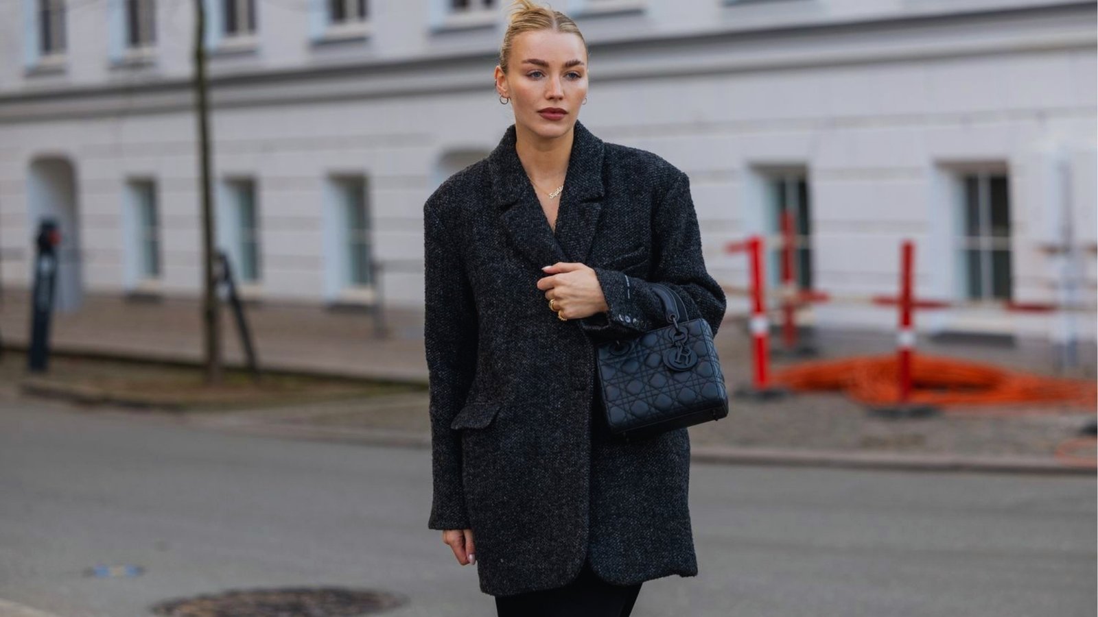 A woman in a black coat and holding a black handbag walks on a city street. The background features a white building and some red and orange construction markers.