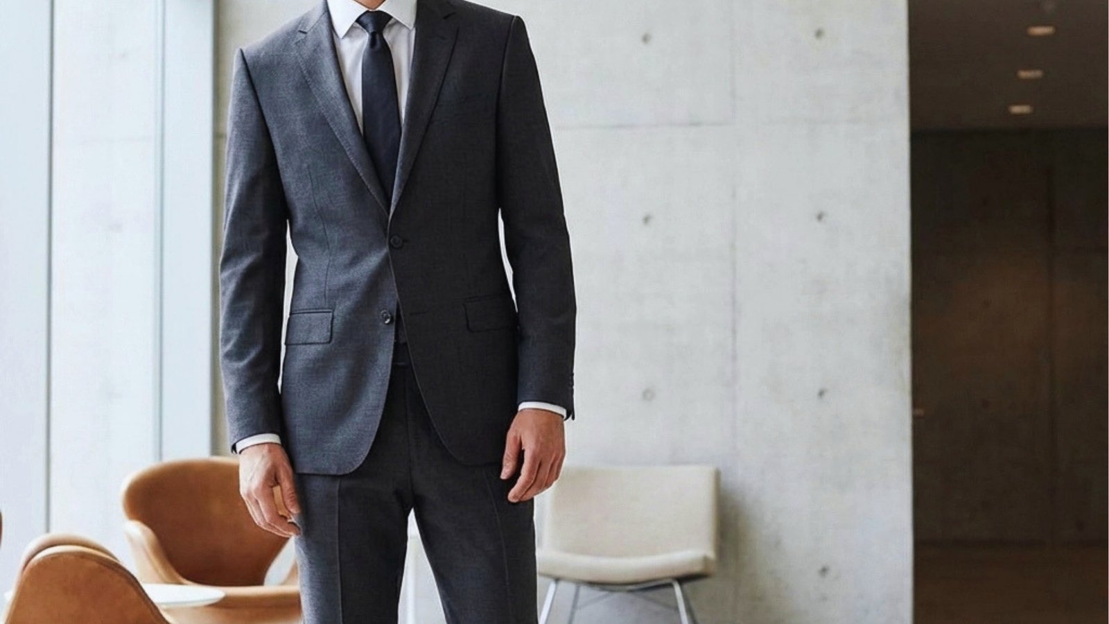 A man wearing a dark gray suit with a white shirt and navy tie stands in a modern, minimalistic office. Neutral tones and soft lighting convey professionalism.