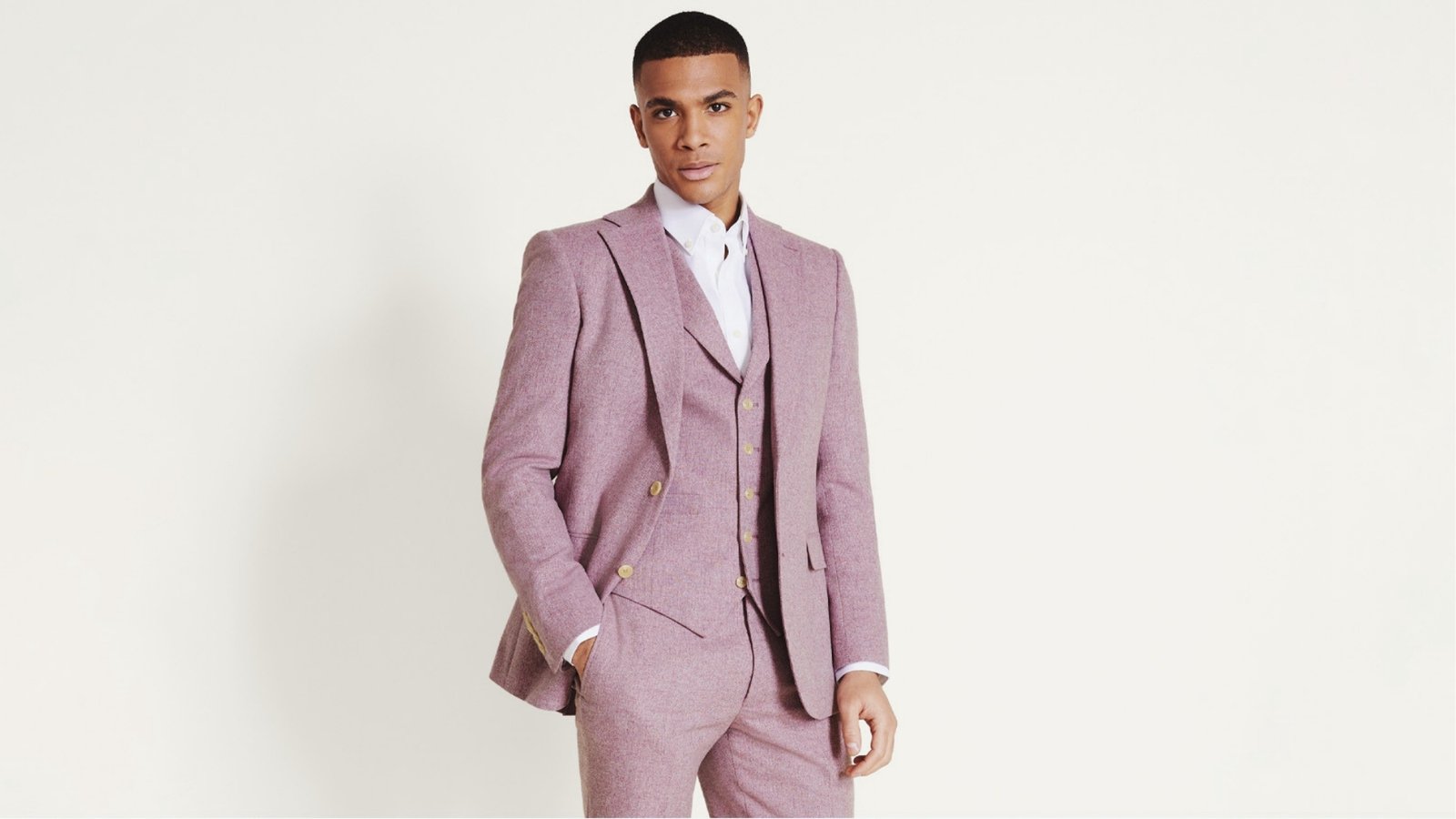 A person confidently wears a tailored pink three-piece suit with a white shirt. The setting is minimalist, and the tone is sophisticated and stylish.
