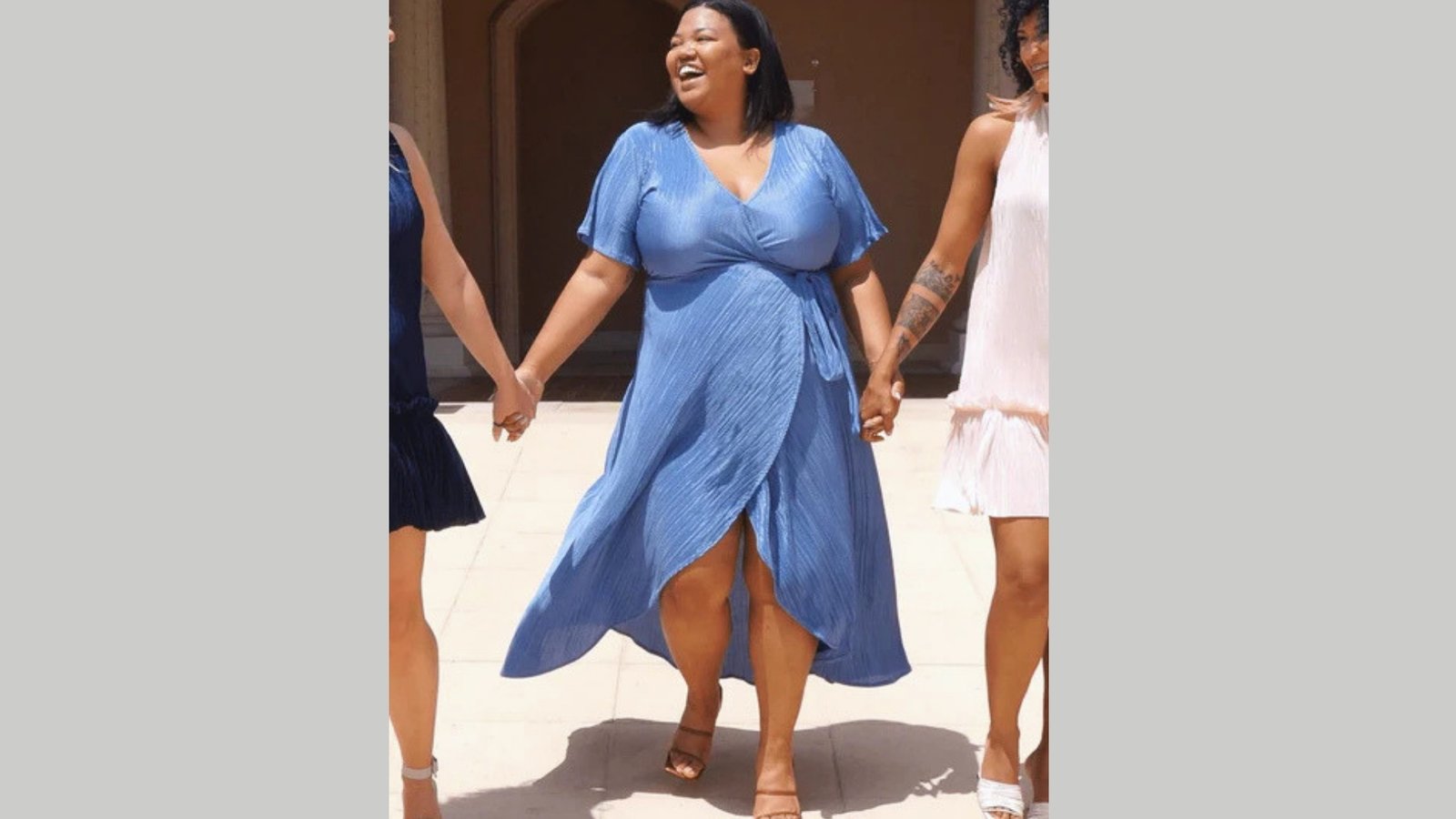 A woman in a blue dress, smiling and laughing, holds hands with two others. They walk outdoors on a sunny day, exuding happiness and camaraderie.