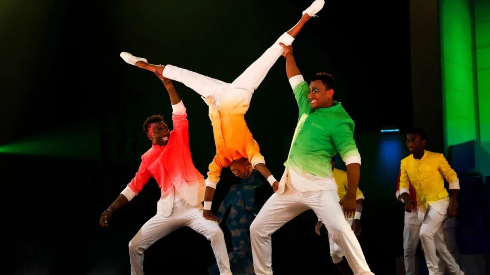 Three performers in colorful costumes, one in a handstand supported by two others, express joy and energy on a dimly lit stage.