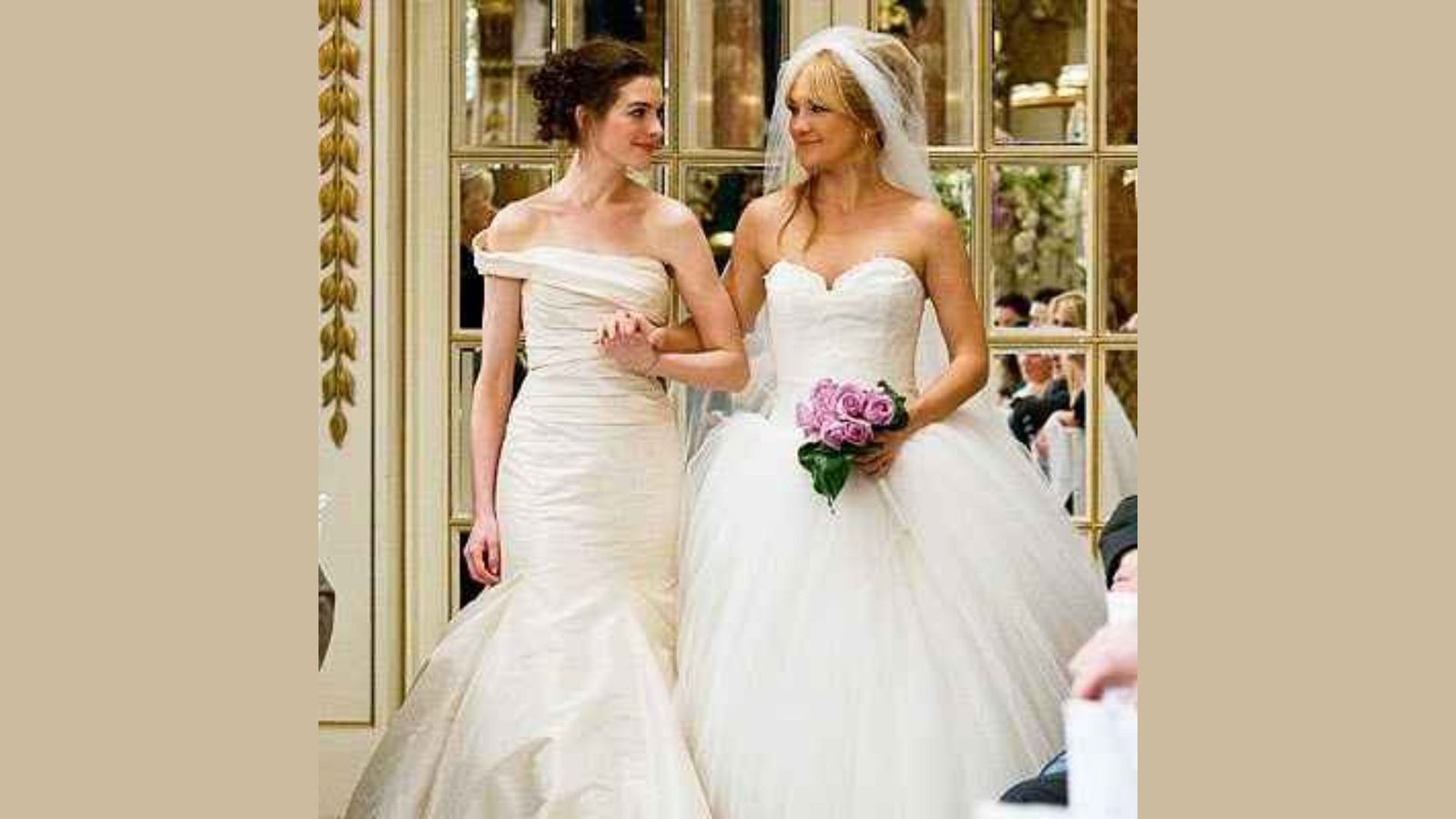 Two brides in elegant gowns walk arm-in-arm, exchanging joyful smiles. One holds a bouquet of pink roses. They stand in a warmly lit, ornate room.