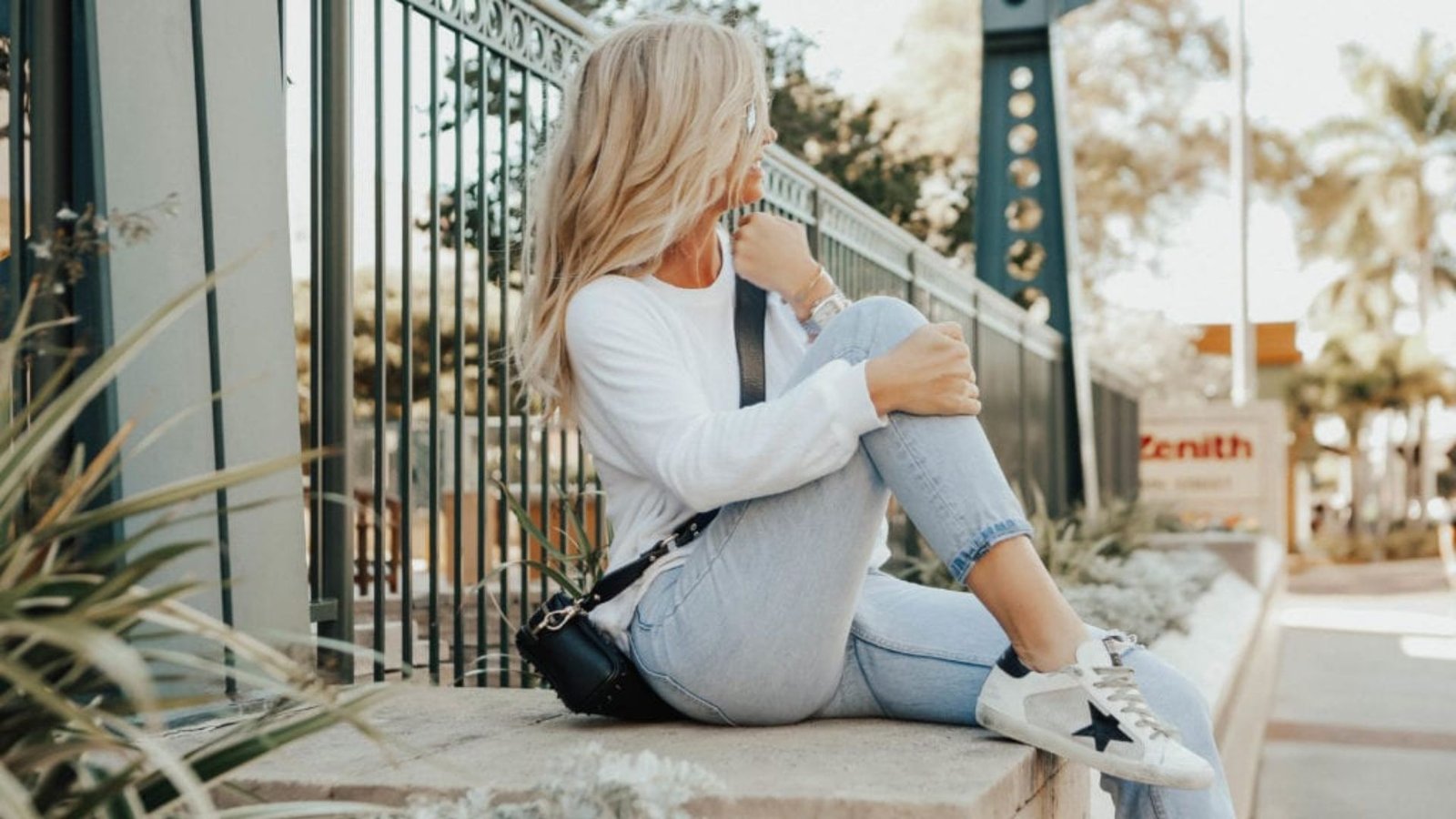 Blonde woman sitting outdoors in a casual mom outfit with a white long-sleeve tee, cuffed light wash jeans, white sneakers with a black star, and a black crossbody bag.