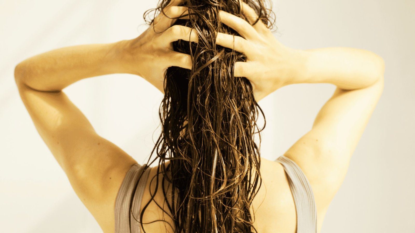 A woman with long hair stands in front of a white wall, demonstrating a hair care technique for scalp massage.
