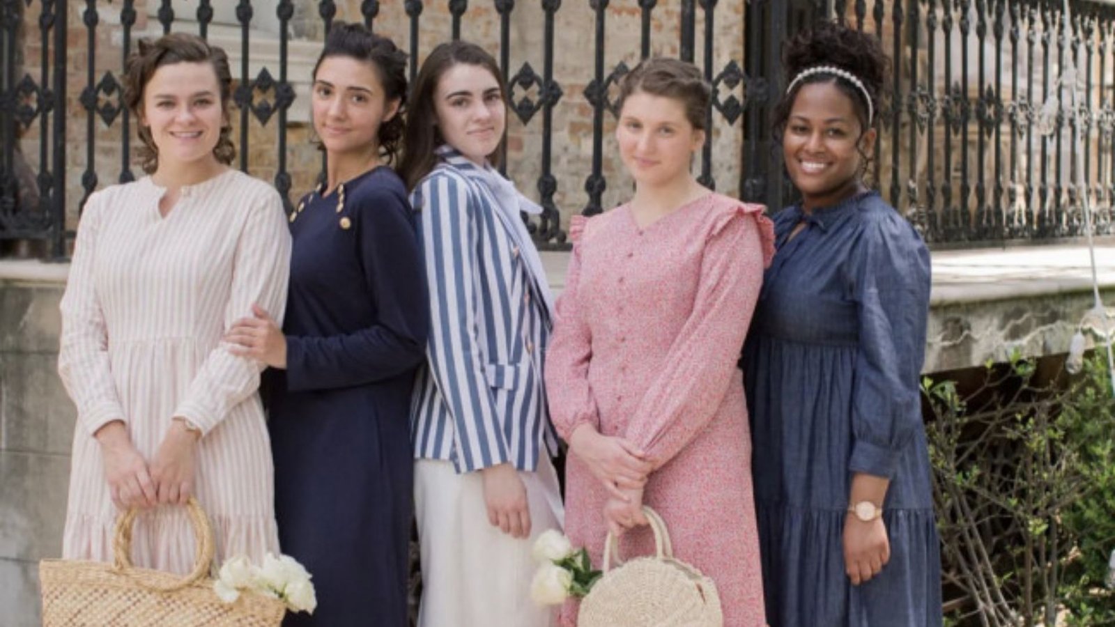 Five women stand in a row, wearing vintage-style dresses in various colors and patterns, holding woven handbags. They are smiling gently, exuding warmth and camaraderie against a wrought iron fence backdrop.