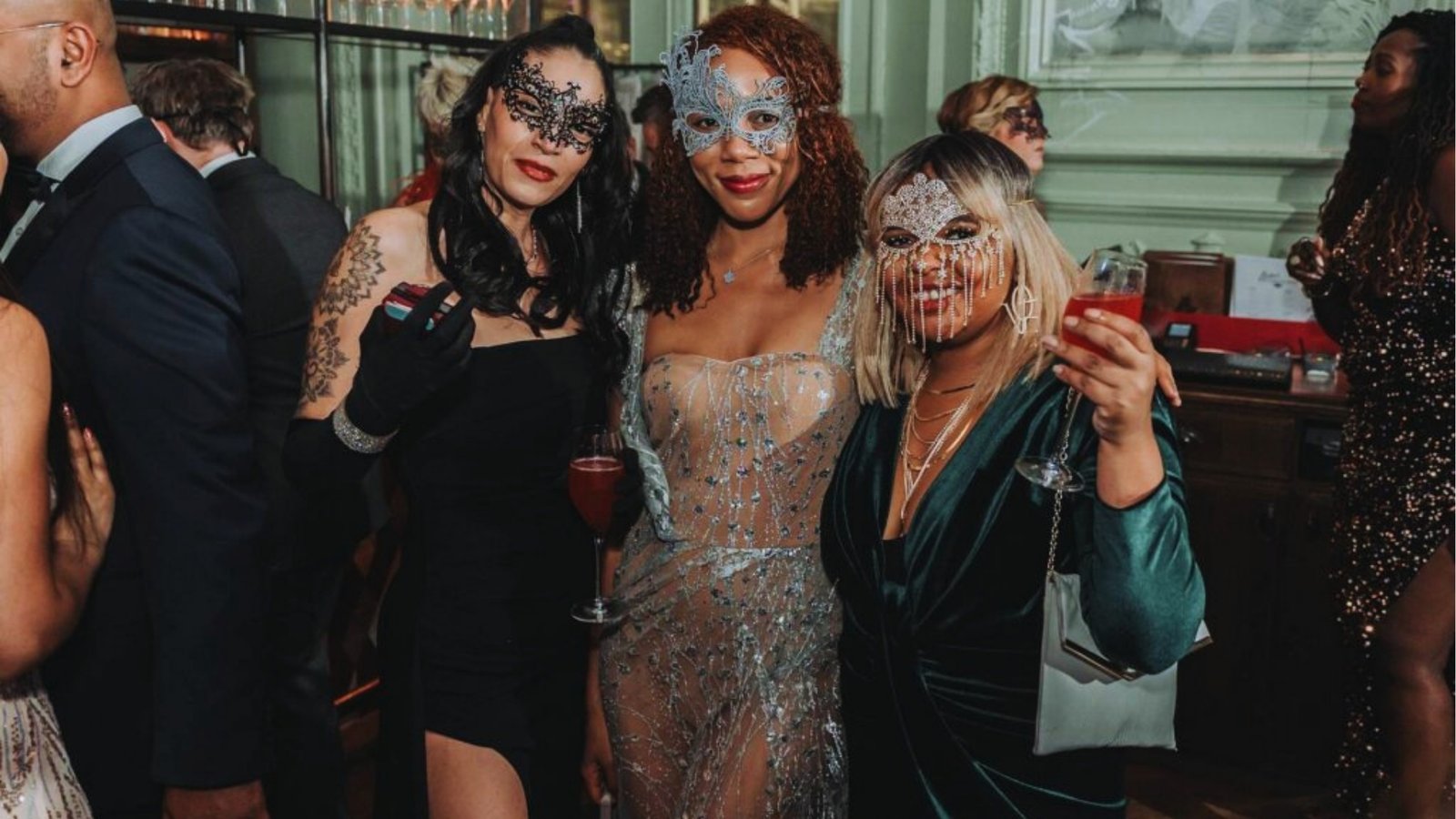 Three women in elegant dresses and ornate masks enjoy a masquerade ball, holding cocktails and smiling. The atmosphere is festive and glamorous.