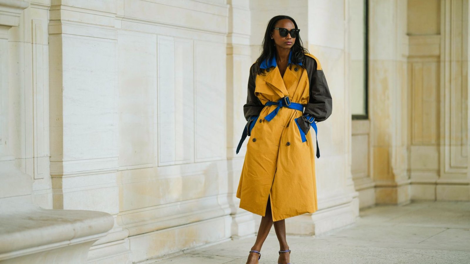 A person stands confidently against a cream-colored wall wearing a yellow coat with blue accents, stylish sunglasses, and strappy heels, exuding urban chic.