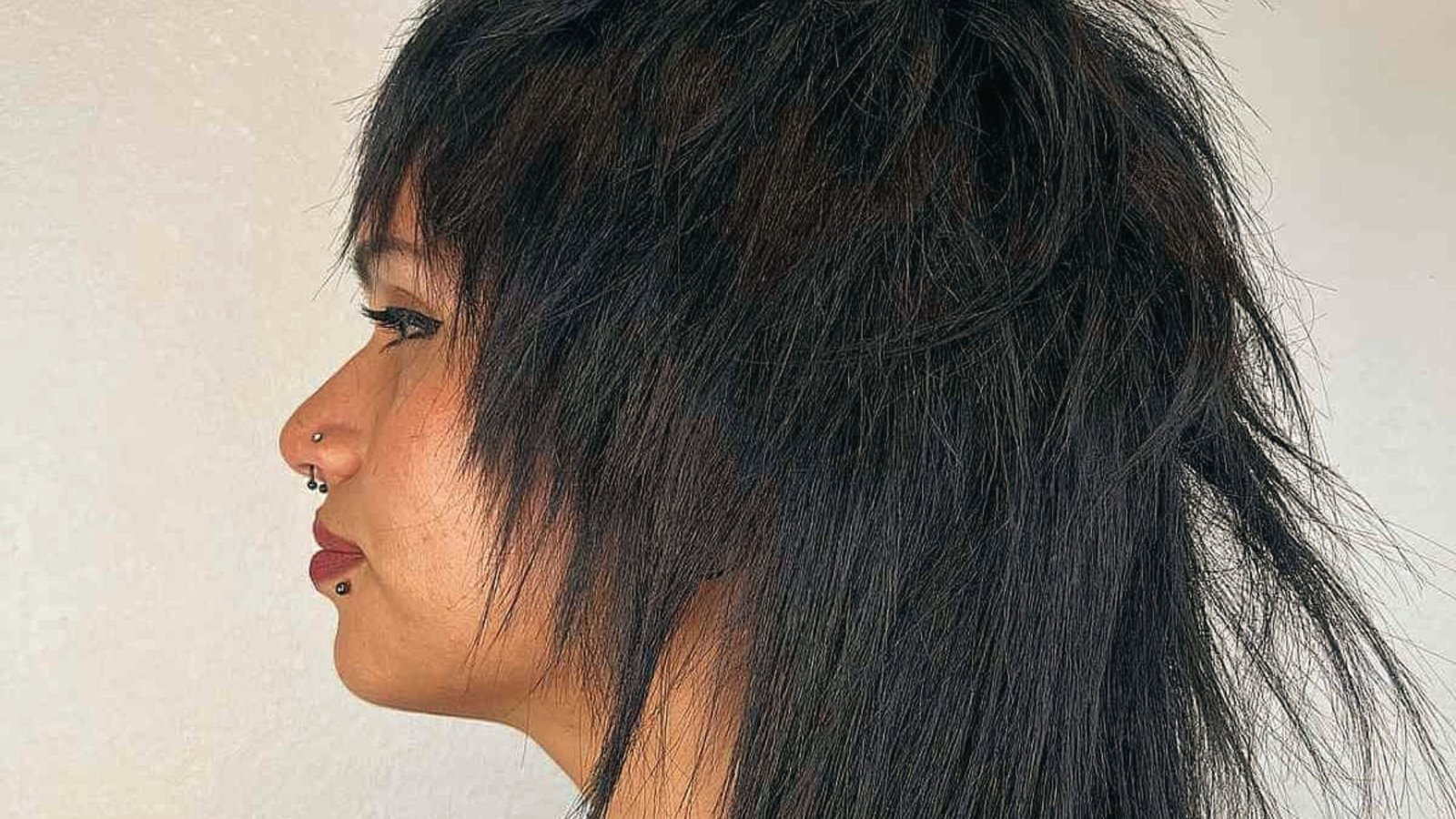 A woman with long hair styled in a disconnected mullet, featuring a facial piercing, gazes confidently at the camera.