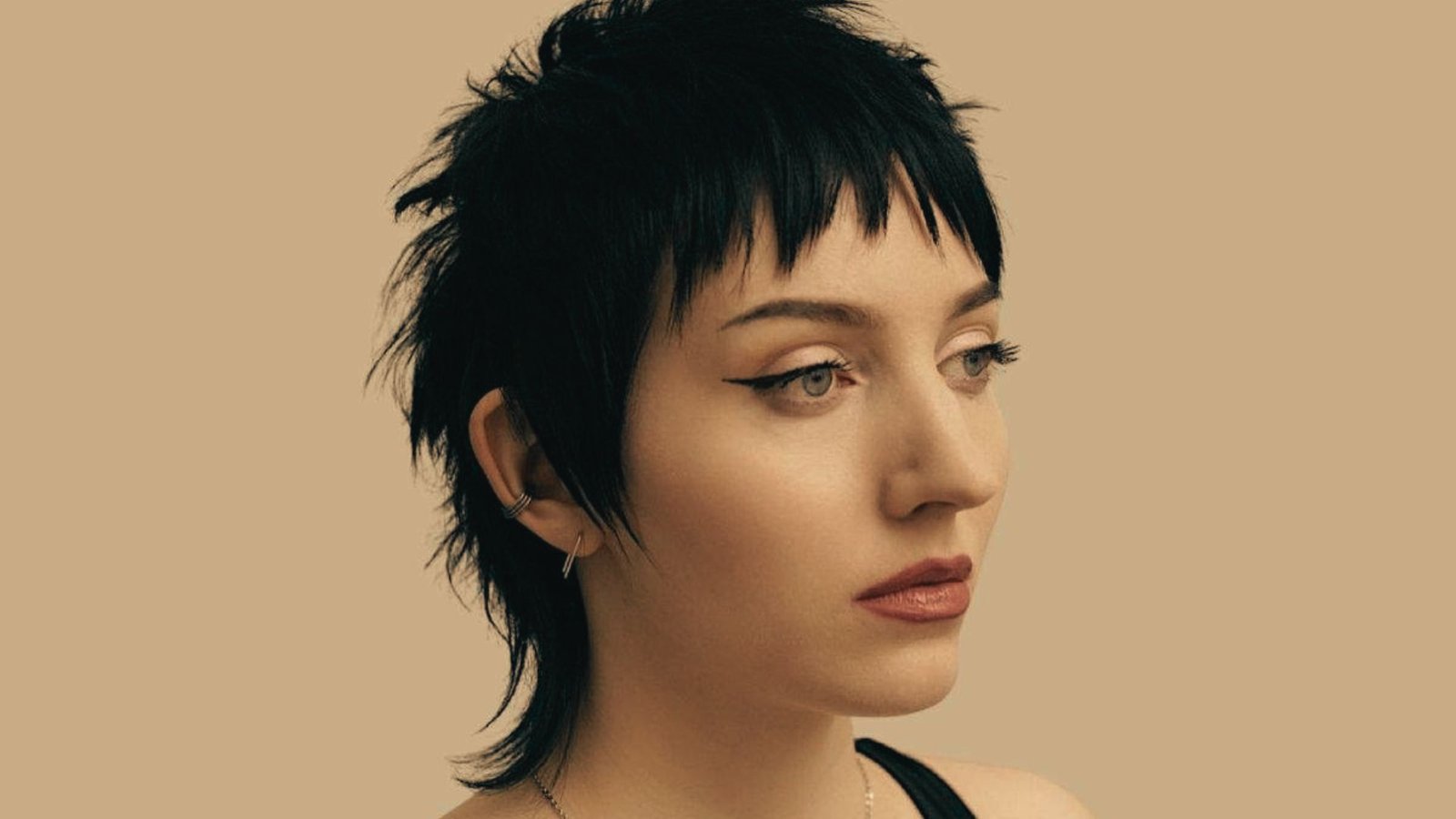 A woman with a wispy mullet haircut, black hair, and a facial piercing, looking confidently at the camera