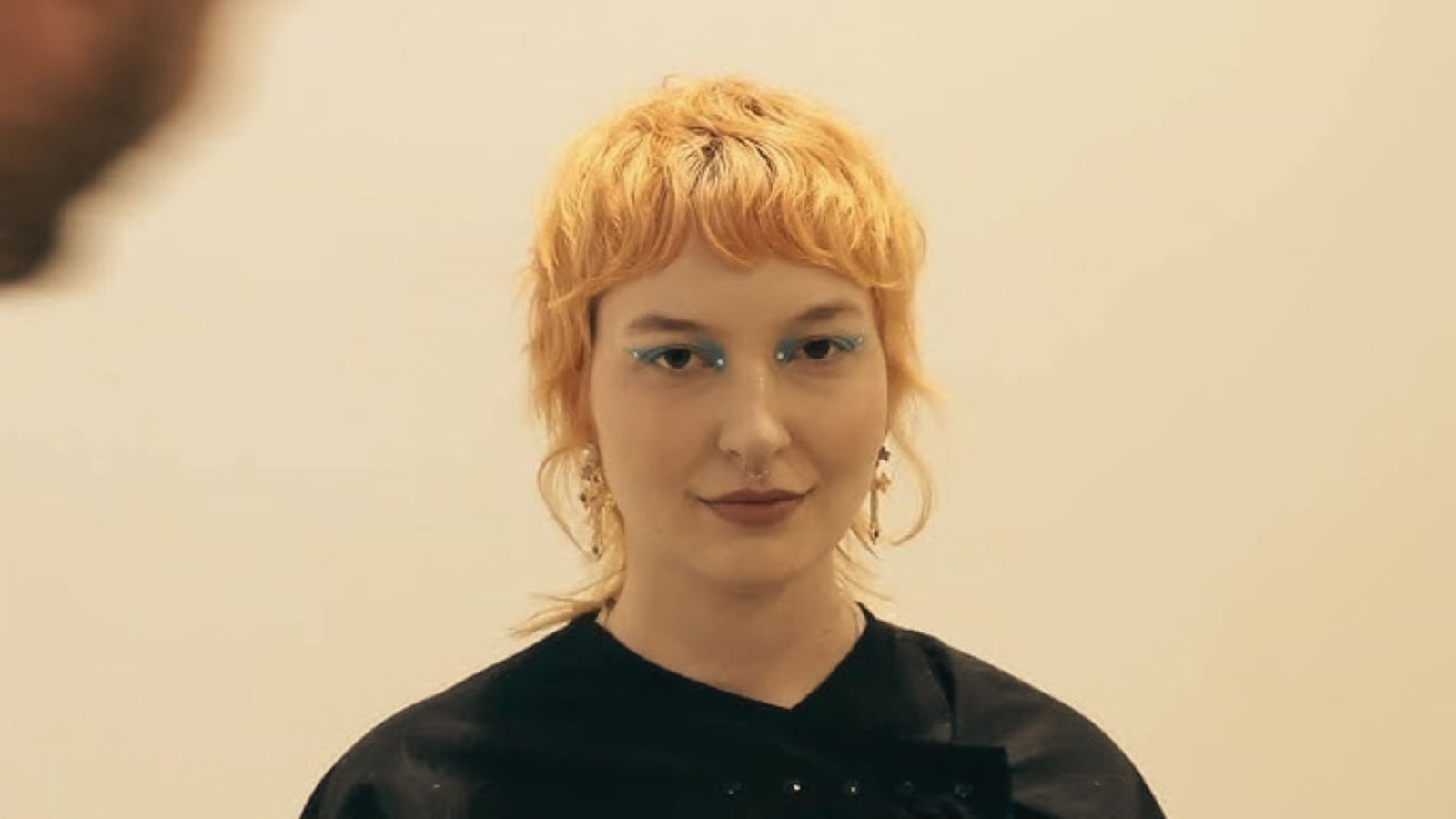 A woman with an orange pixie mullet haircut wearing a black shirt, looking confidently at the camera.