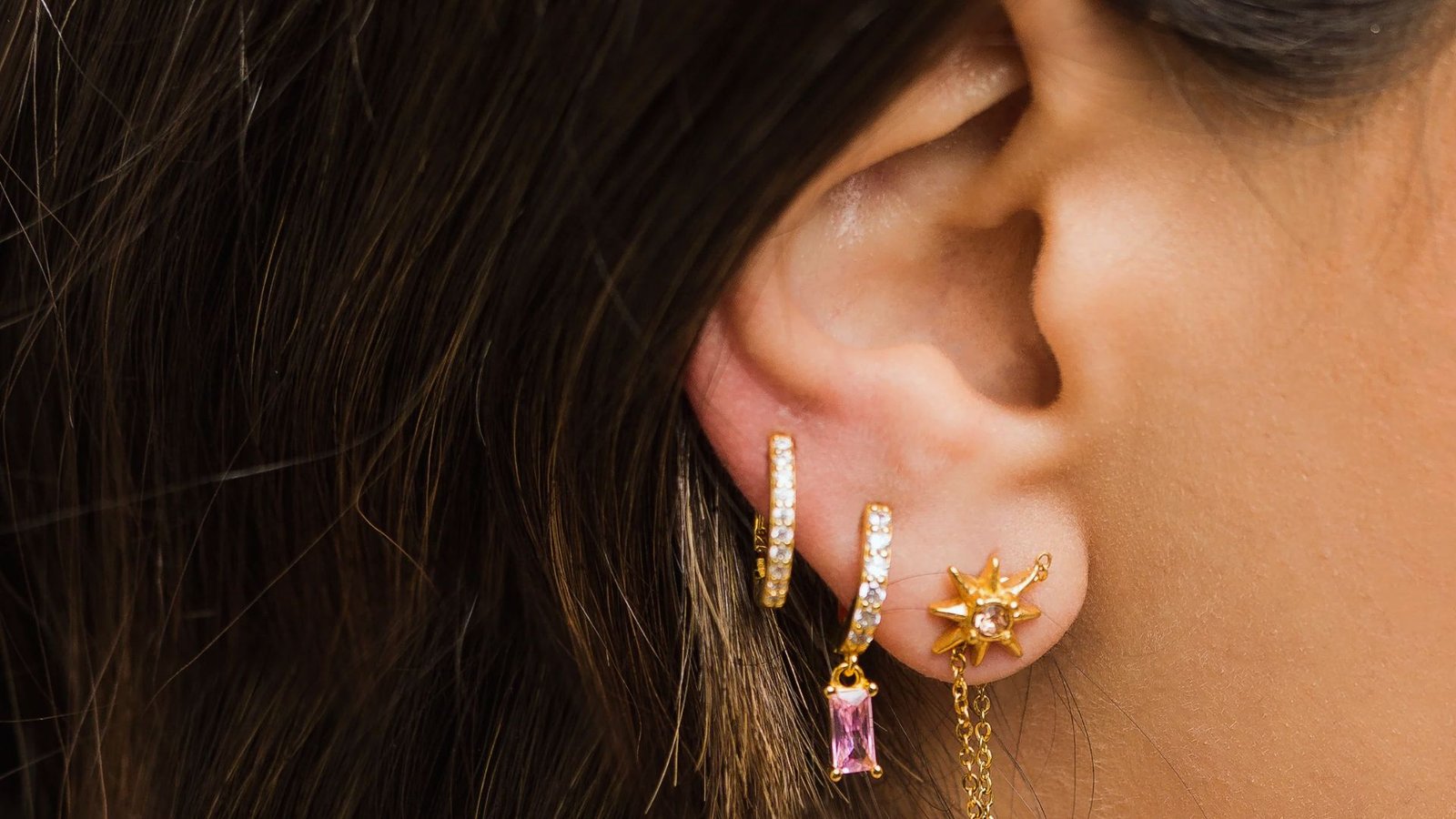 A close-up of a woman's ear wearing three earrings: two gold hoops with small gemstones, and a gold sun-shaped stud with dangling chain and pink gem.