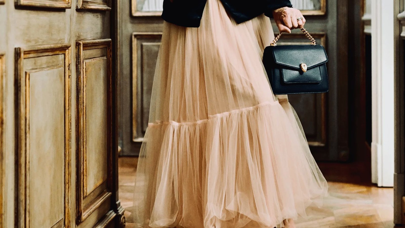 A person in a flowing beige tulle skirt and black jacket holds a stylish black handbag with a chain strap. The setting is a classical, ornate hallway.