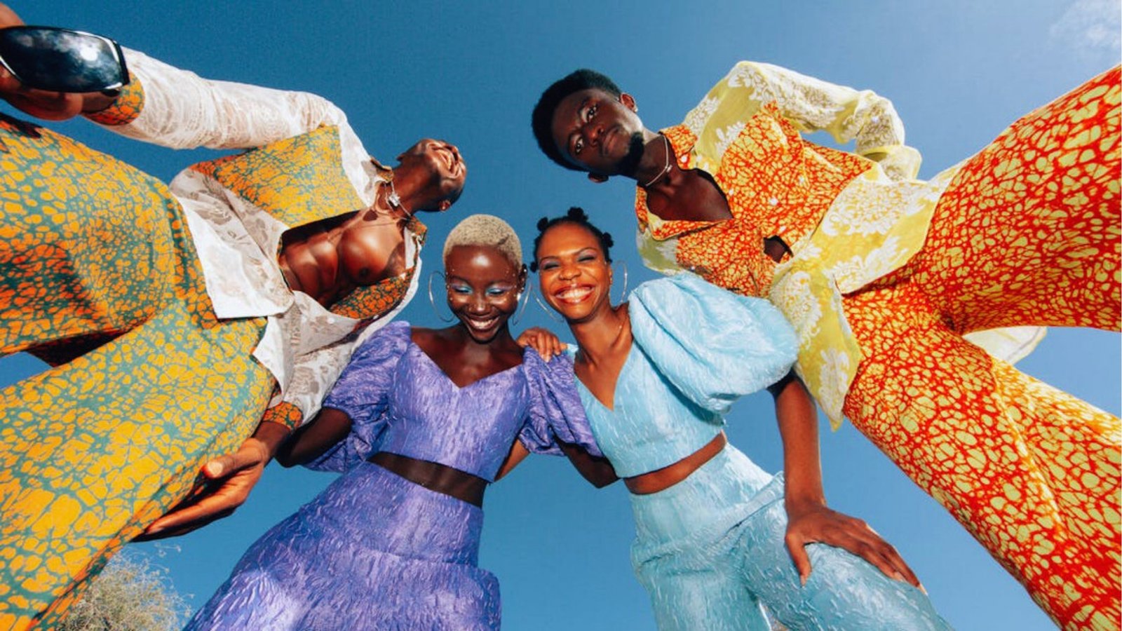 Four people in vibrant, patterned outfits stand close together under a clear blue sky, smiling warmly at the camera, conveying joy and unity.