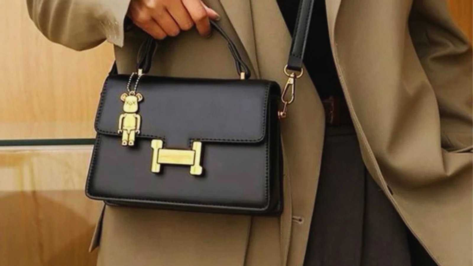 A person in a tan coat holds a stylish black handbag featuring gold accents and a small bear charm, evoking a sense of elegance and sophistication.