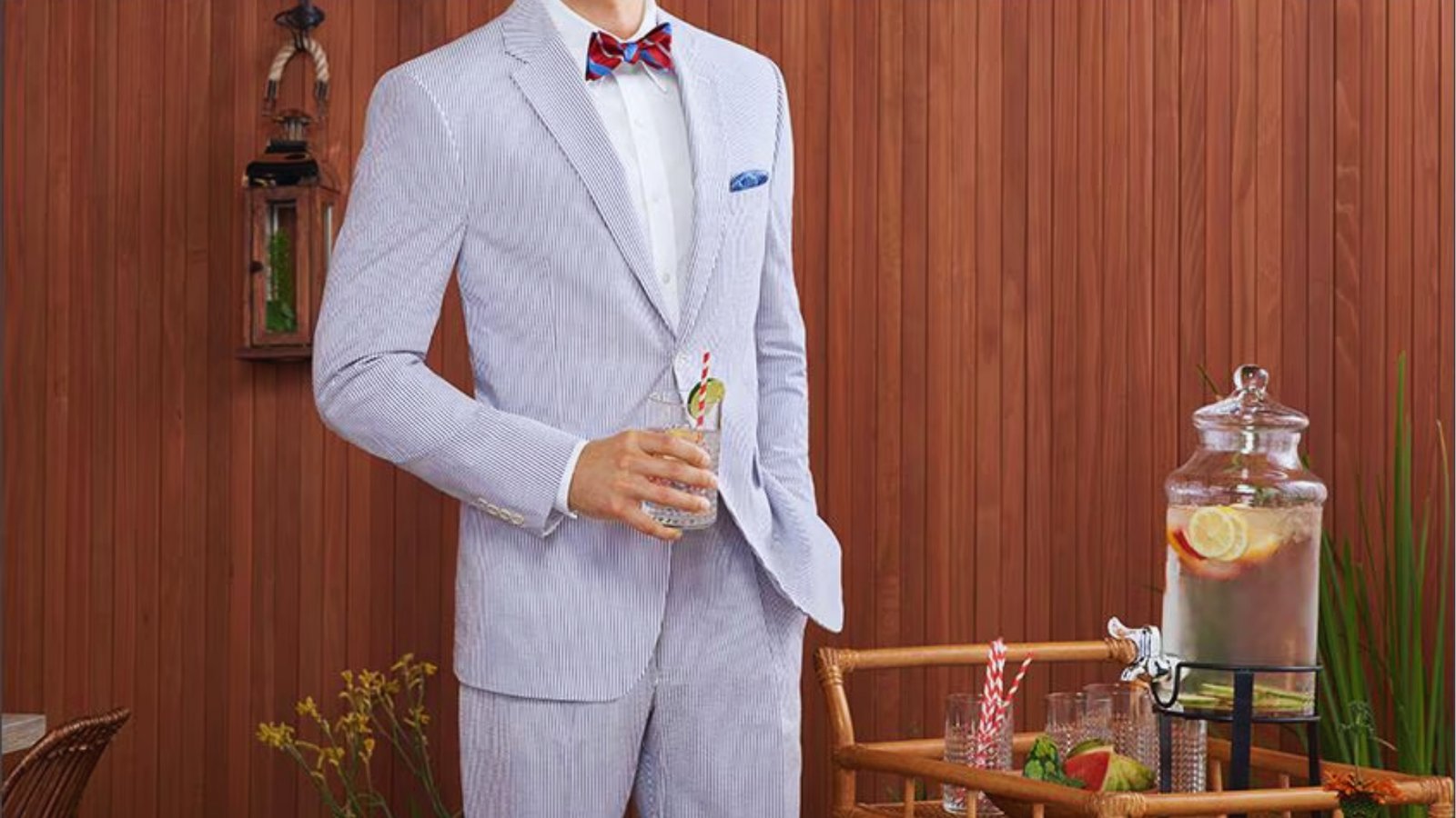 A man in a light-striped suit with a red bow tie holds a drink by a drink cart with infused water and glasses. The wooden backdrop adds warmth.