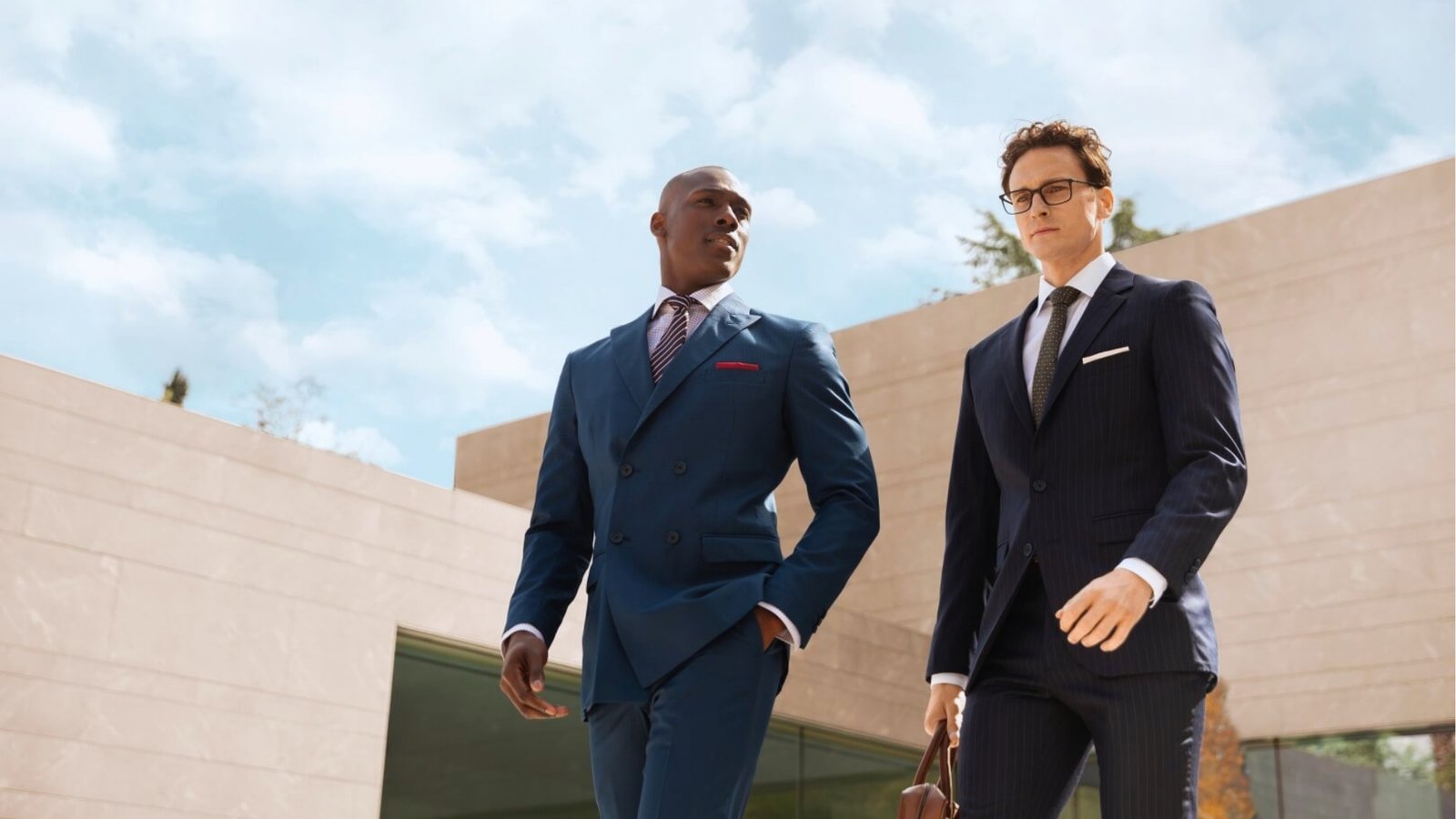 Two men in sharp suits walk confidently outdoors in front of a modern building under a clear blue sky. They convey professionalism and purpose.