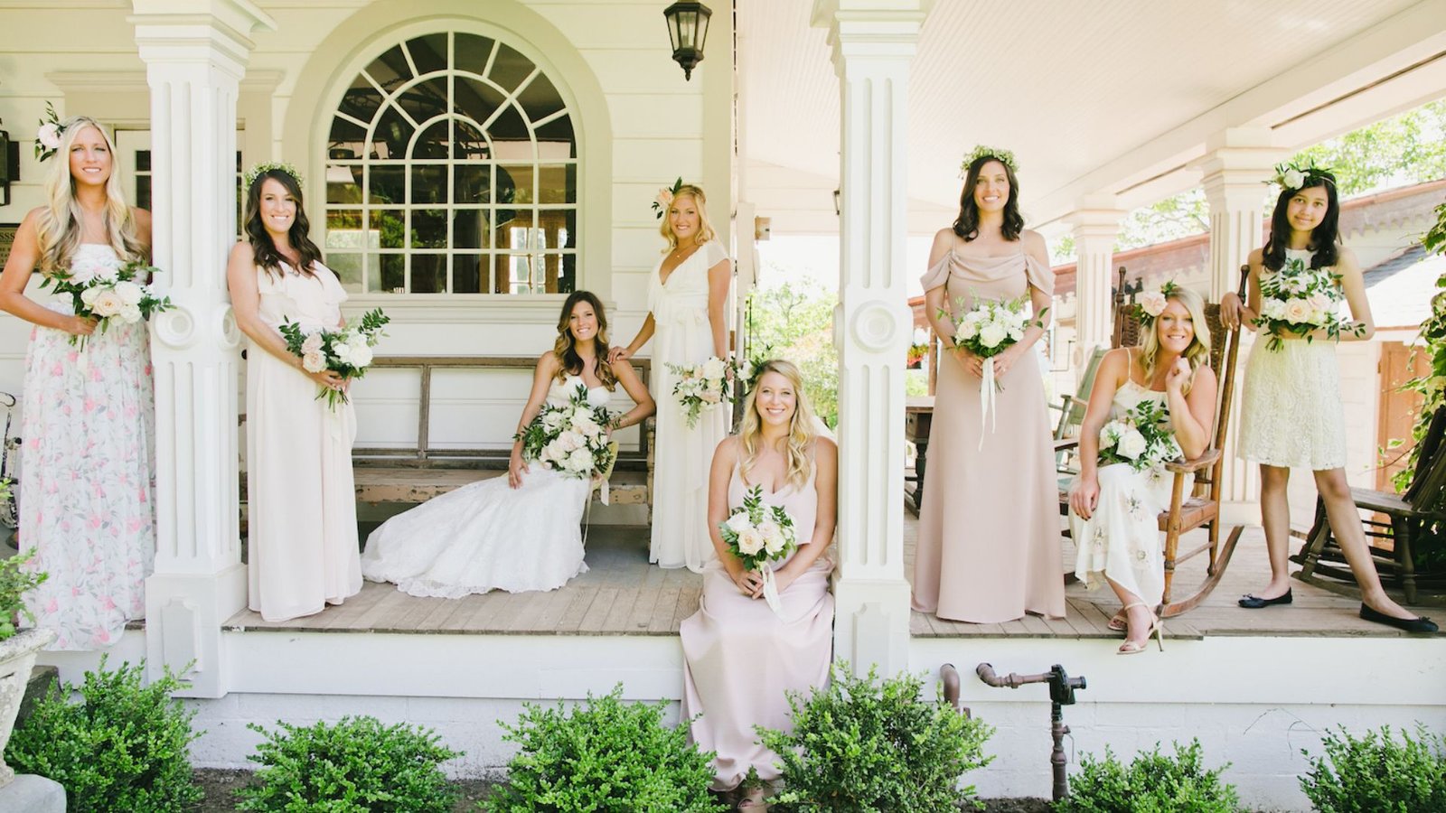 Seven bridesmaids in pastel dresses and one woman in a wedding gown pose with bouquets on a charming porch, conveying joy and elegance.