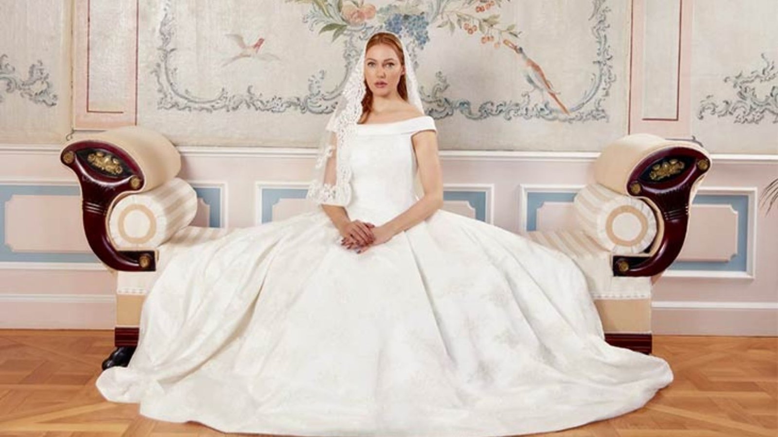 A bride in an elegant white gown sits on an ornate, vintage chaise longue. The backdrop features pastel floral and bird motifs, creating a serene, classic atmosphere.