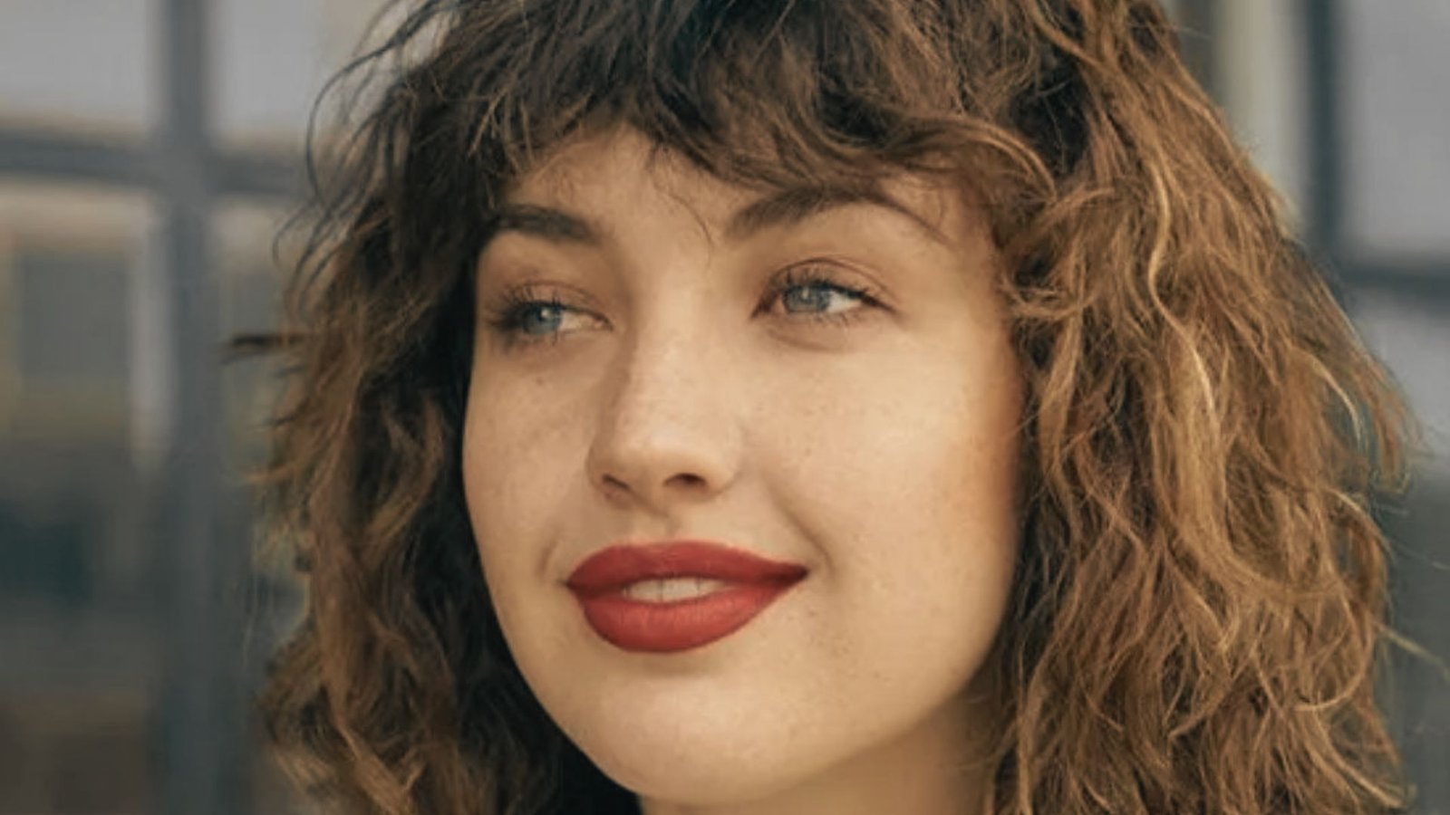 A woman with a round face, curly hair styled with curtain bangs, and wearing bold red lipstick.