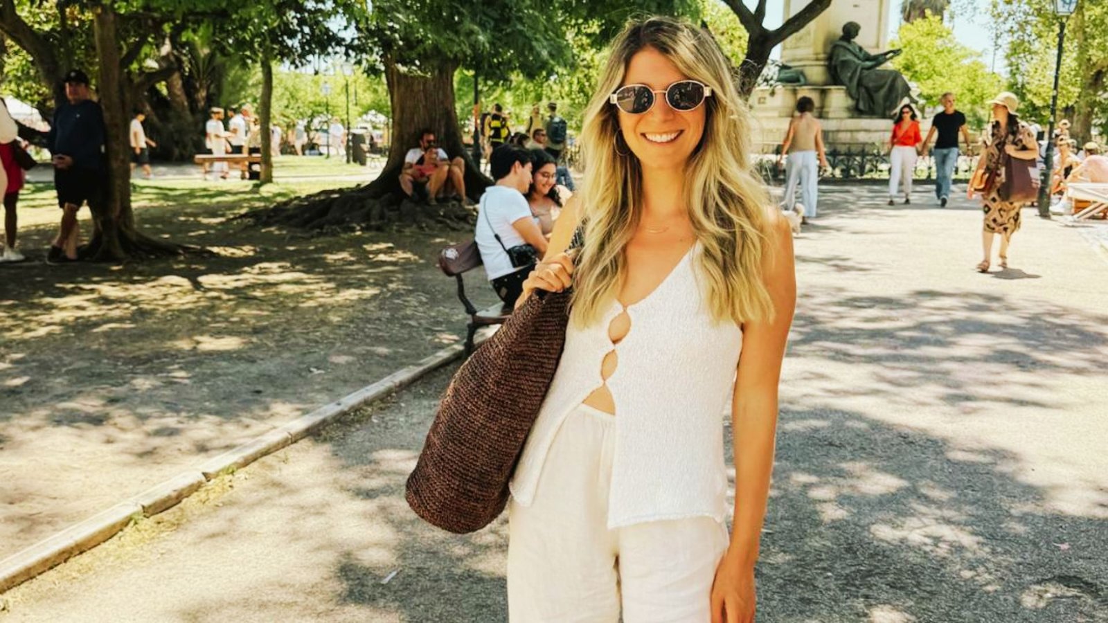 A woman in white pants and sunglasses holds a brown bag, embodying effortless mom fashion with a stylish yet relaxed look.