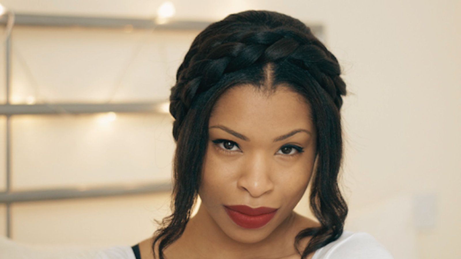 A woman with milkmaid braids and bangs, wearing bold red lipstick, smiles confidently at the camera.