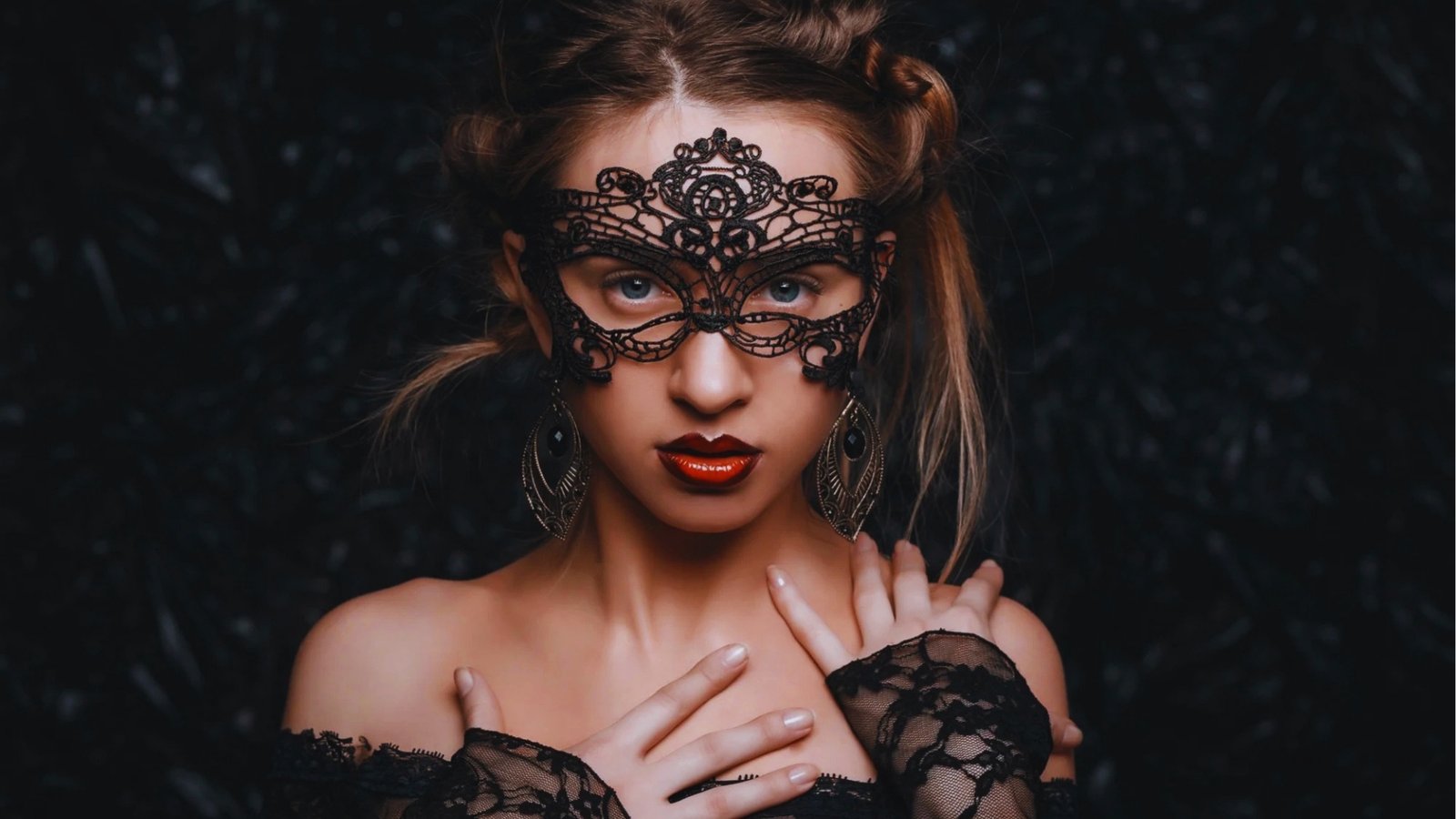 A woman wearing an intricate black lace mask gazes directly at the camera. She has bright red lips, silver earrings, and is dressed in lace, exuding mystery.