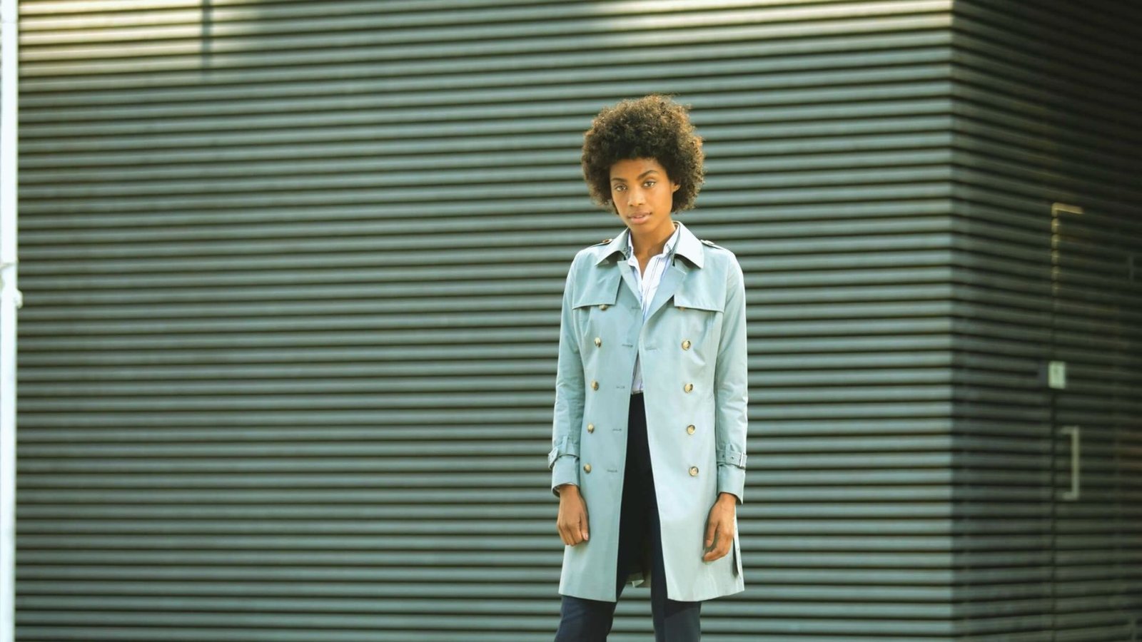 A person with curly hair stands confidently in front of a striped metal wall, wearing a light trench coat and dark pants. The tone is modern and poised.