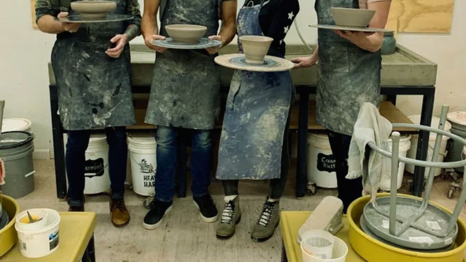 Four people in aprons hold pottery pieces on wheels in a studio. The setting is creative and hands-on, with various clay tools and equipment visible.