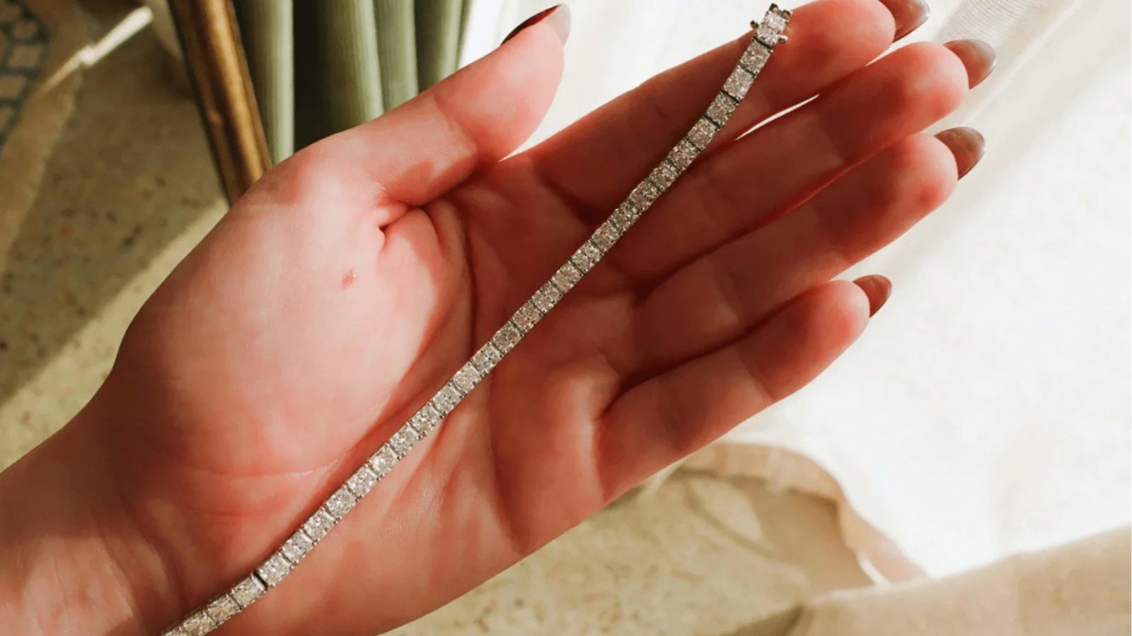 A hand holds a sparkling diamond bracelet, with sunlight enhancing the glimmer. The setting is soft and elegant, conveying a sense of luxury and beauty.