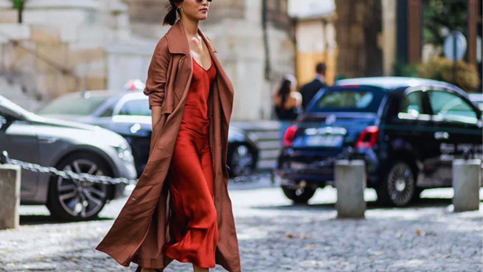 A woman in a flowing red dress and brown trench coat walks confidently on a city street, surrounded by parked cars, exuding style and elegance.