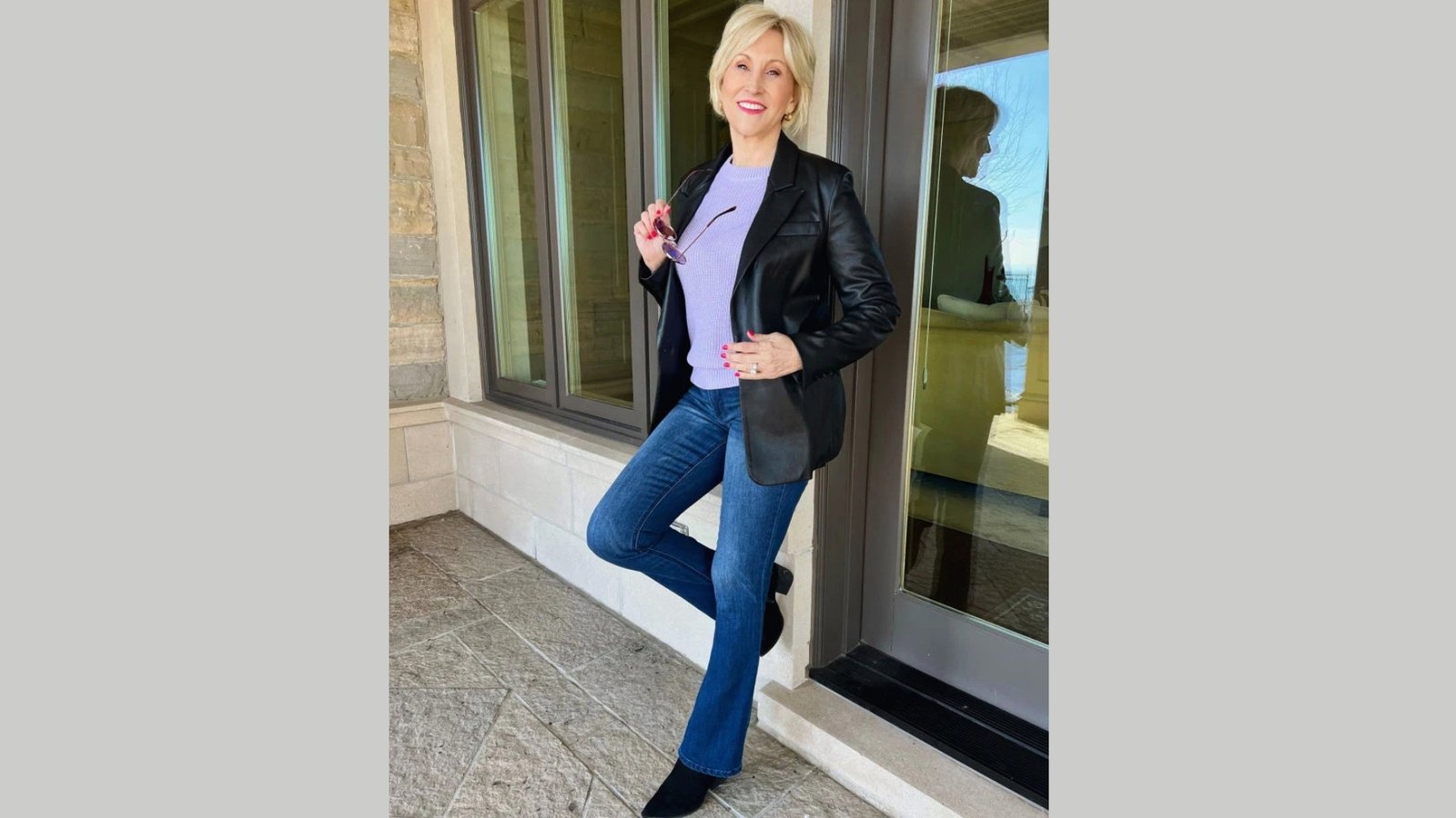A person stands confidently against a glass door, wearing a black leather jacket, purple sweater, and jeans, holding sunglasses, exuding a relaxed, stylish vibe.