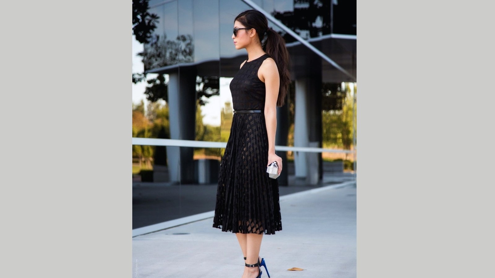 Woman standing sideways, wearing a sleeveless black dress and sunglasses, holding a clutch. She exudes elegance in an urban setting with reflective glass in the background.