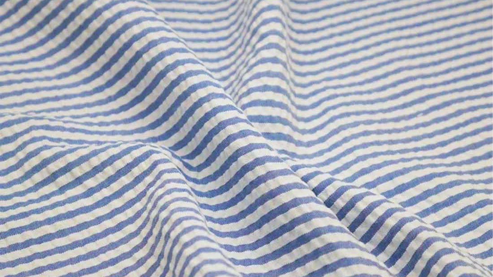 Close-up of a blue and white striped seersucker fabric, with soft waves and folds creating a textured, airy feel. The tone is light and summery.