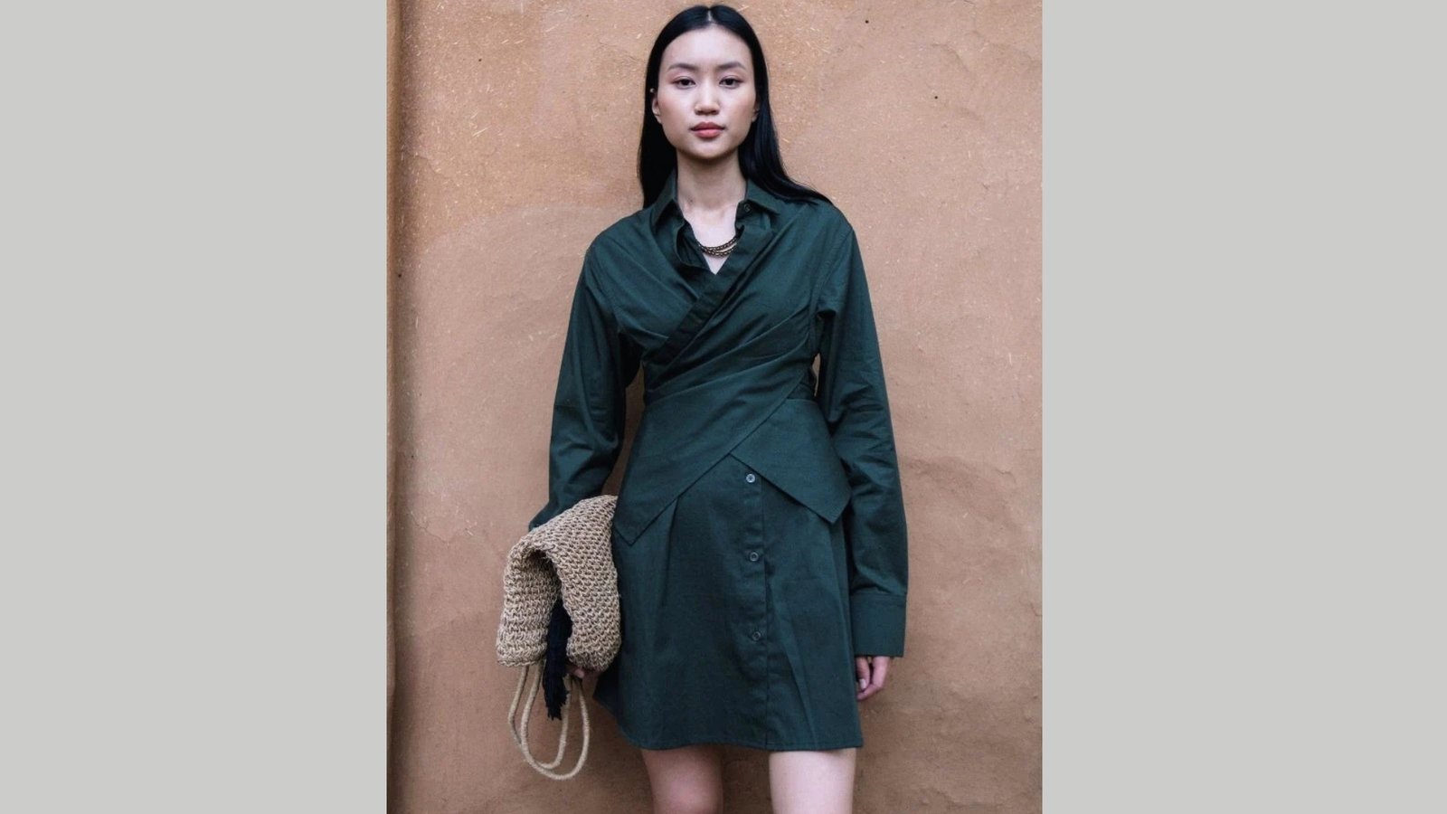A woman stands against a textured, earthy wall wearing a dark green, buttoned dress. She holds a woven bag, exuding a calm and confident aura.