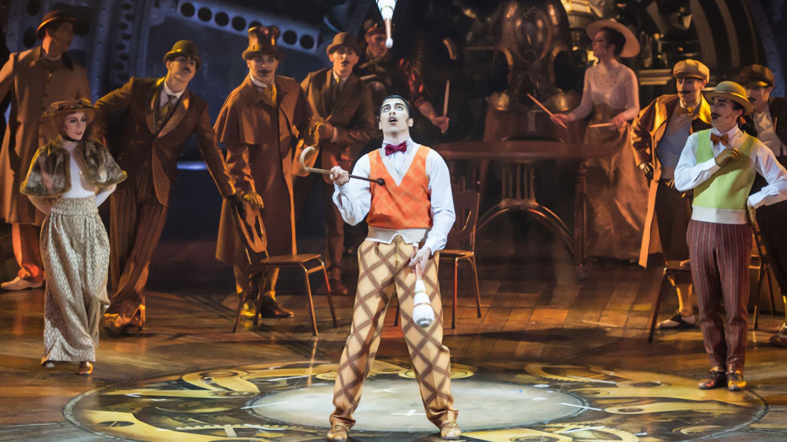 A performer in an orange vest and checkered pants juggles on a vibrant stage. He's surrounded by a lively ensemble in vintage attire, creating an energetic, theatrical atmosphere.
