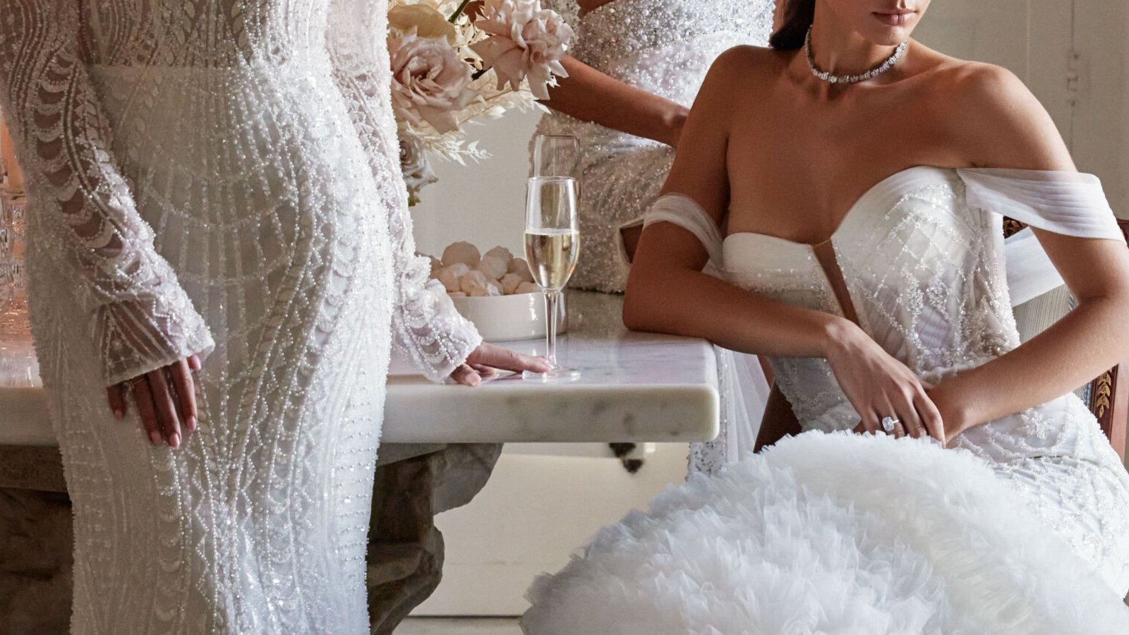 Elegant scene of three women in embellished white gowns around a marble table with a champagne glass and floral centerpiece. The tone is luxurious.